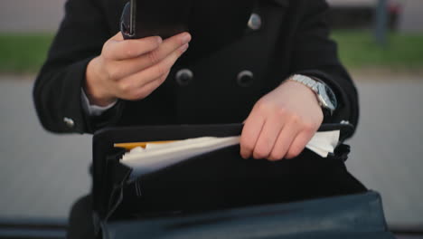 close up of individual hand in black clothing going through documents in bag, retrieving black diary, background featuring greenery, unclear figure, interlocked path in serene outdoor environment