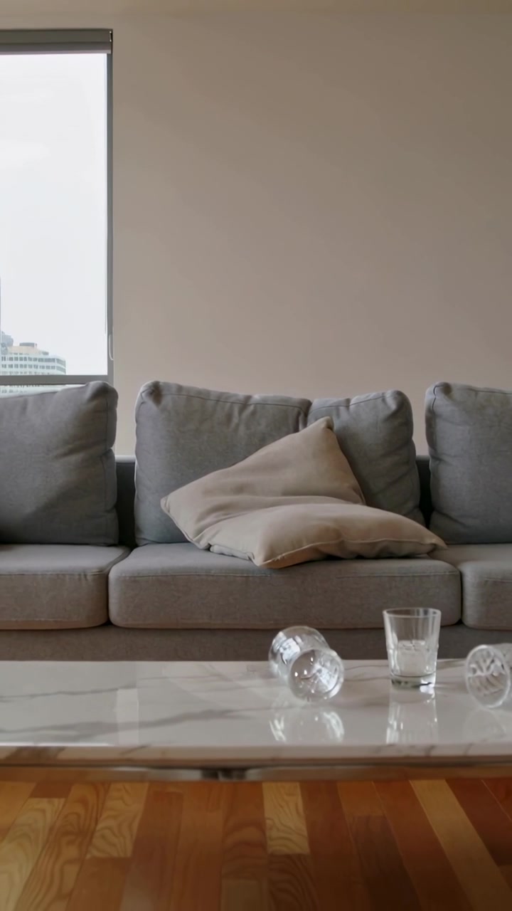 Vertikal video: Rullende klart glas fra spidsen hen over bordet i en lejlighed, lander nær en grå sofa