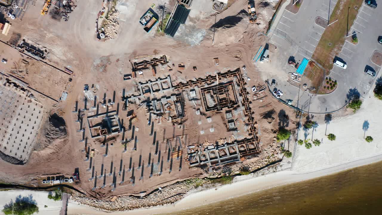 Pantai berpasir putih dengan pohon palem yang tersebar di sepanjang perairan dangkal, sementara tempat parkir terdekat dan lokasi konstruksi yang berdekatan menunjukkan pembangunan perkotaan di lingkungan pesisir.