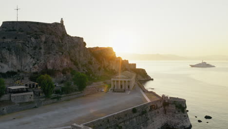 Saint George Church in Old Fortress of Corfu at sunrise with calm sea below, tracking left backlit sun on mountains