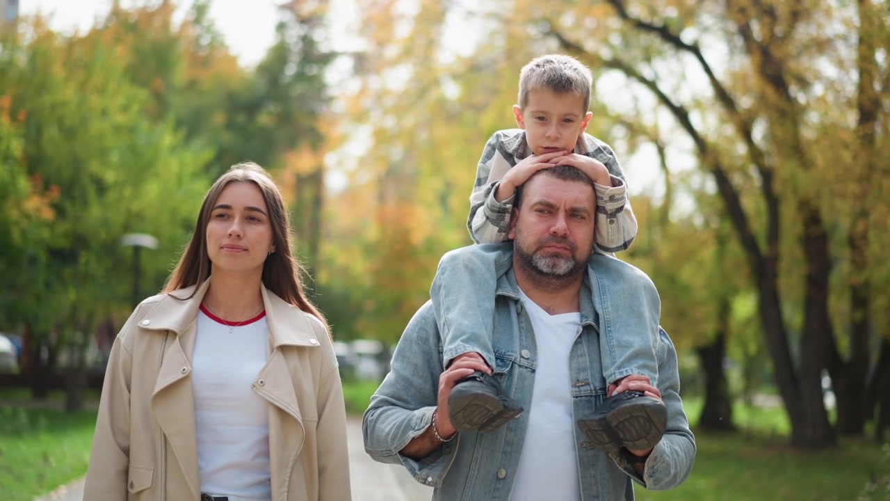Primo piano di un uomo che porta un bambino piccolo sulle spalle mentre cammina con una donna in un parco all'aperto. La famiglia si muove lungo un sentiero circondato da alberi verdi e una morbida luce naturale che crea un'atmosfera serena.