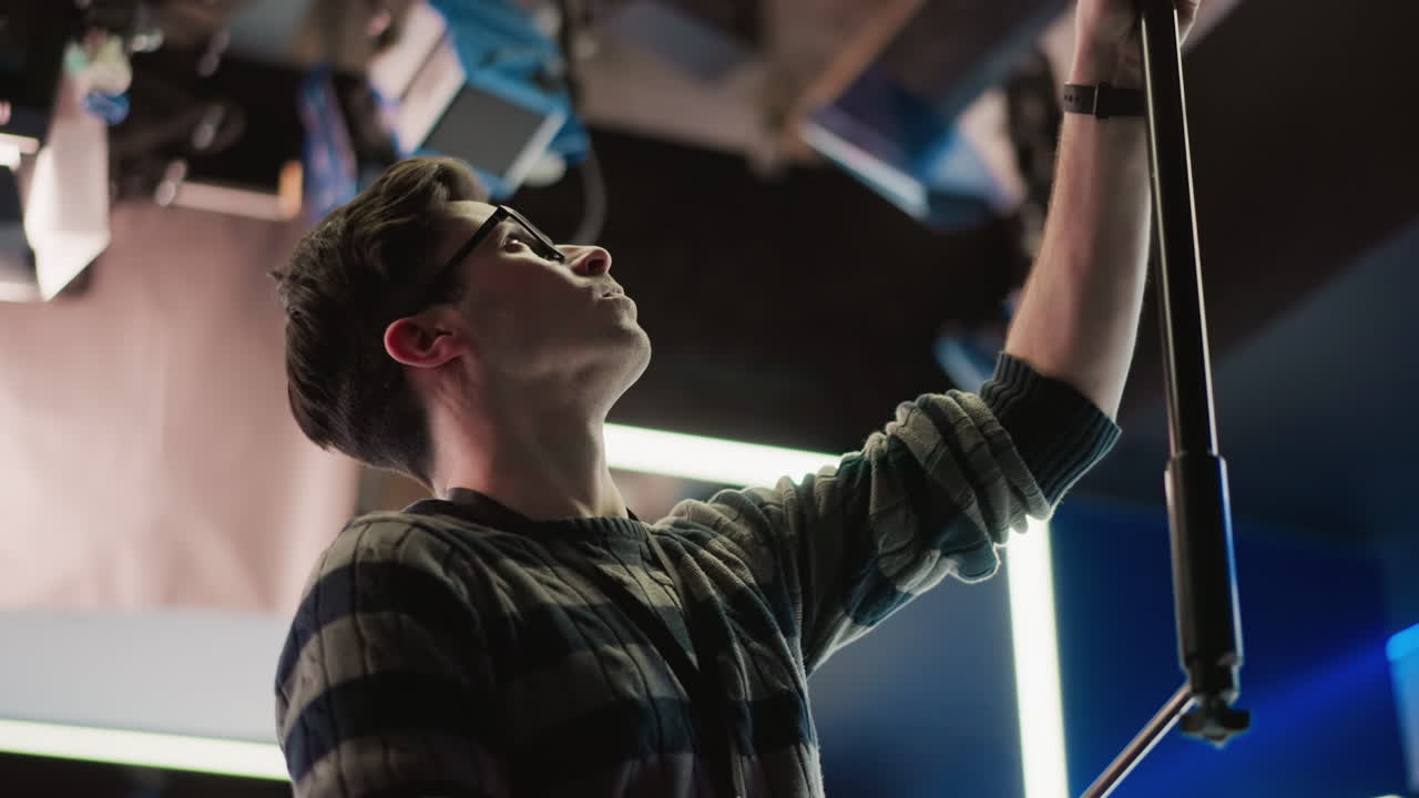 Technician adjusting boom pole in broadcast studio, looking up at lights. Professional media production setup with camera, lighting, and modern studio environment