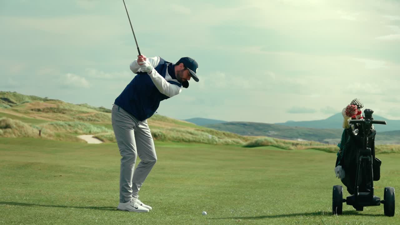 남자 골퍼가 골프 가방 의 완벽한 그림 같은 푸른 하늘의 날에 아일랜드 링크스 골프 코스의 초록색 매니큐어드 페어웨이에서 을 치고 접근합니다.