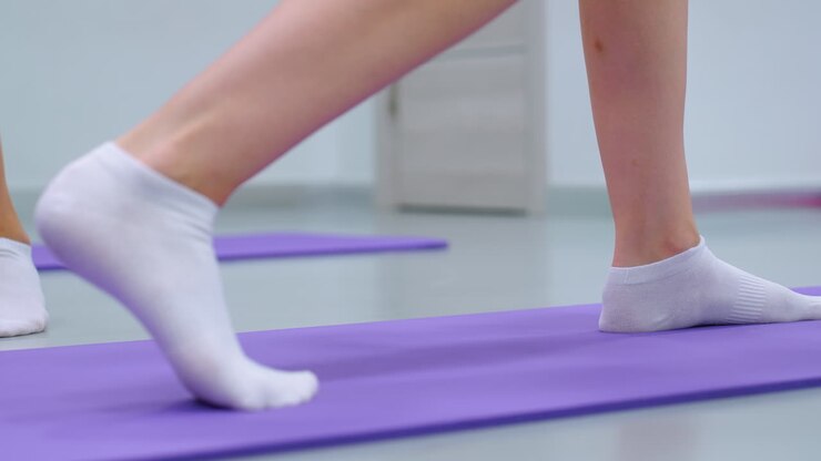 Close-up leg view of a lady in socks lifting a weight on a purple mat, showing dynamic exercise movement with another person partially visible in the background