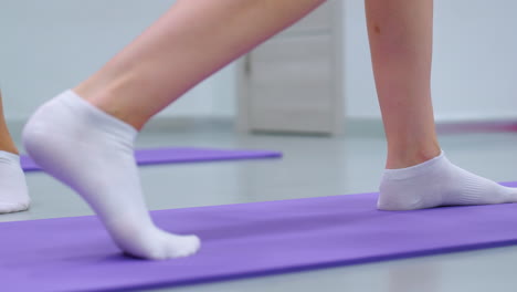 close-up leg view of a lady in socks lifting a weight on a purple mat, showing dynamic exercise movement with another person partially visible in the background