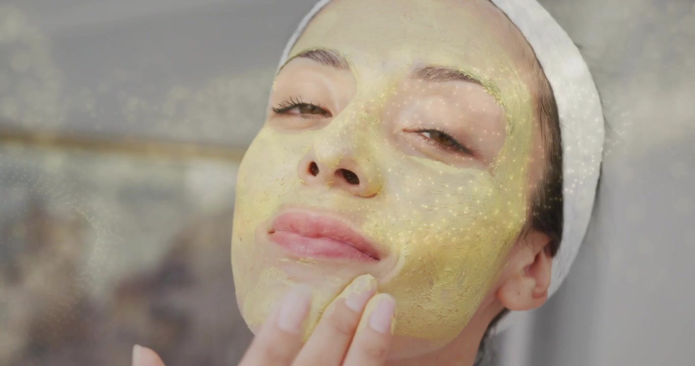 Donna con fascia per capelli che si tocca il mento con la punta delle dita, stendendo una maschera viso dorata per una pelle luminosa.