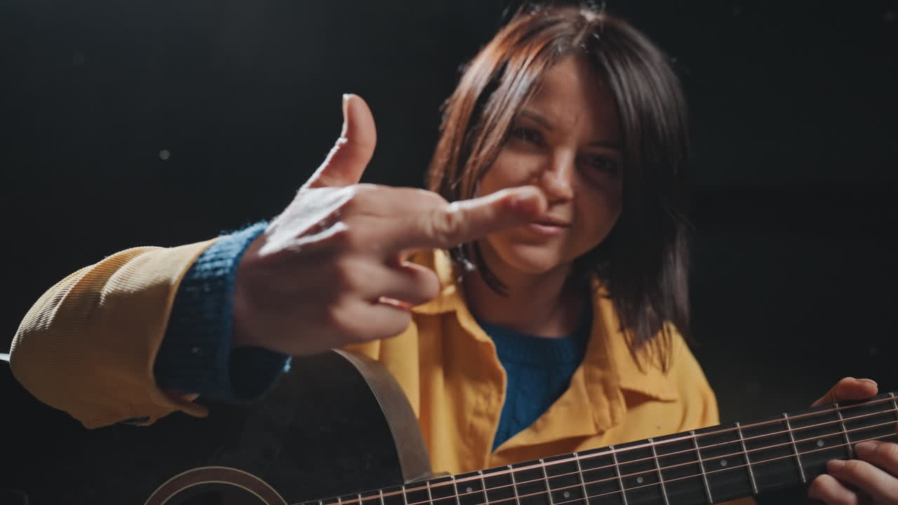 Donna sorridente che suona la chitarra, musicista da campeggio in giacca gialla. Gesto giocoso con le dita ed espressione con la lingua di fuori. Illuminazione da studio spontanea, un'atmosfera accogliente per un canto collettivo con energia folk acustica.