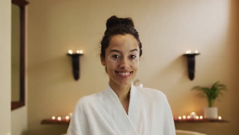 portrait of happy biracial woman in robe standing in bathroom and looking at camera