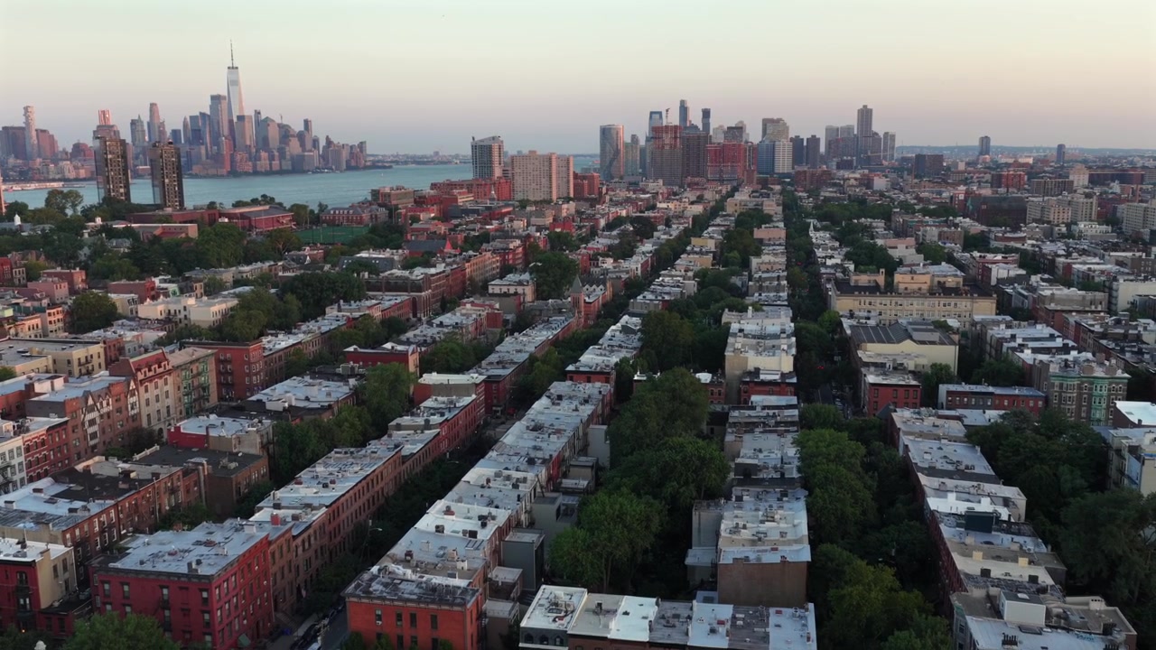 Rækker af brownstone-tage strækker sig gennem beboelsesområdet Hoboken, New Jersey, træbeklædte gader fører mod Hudson-kystlinjen, mens Manhattans skyline gløder ved solopgang. Bredt droneoptagelse baglæns.
