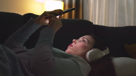 Woman-Wearing-Wireless-Headphones-Lying-On-Sofa-At-Home-At-Night-Streaming-Or-Looking-At-Online-Content-On-Mobile-Phone