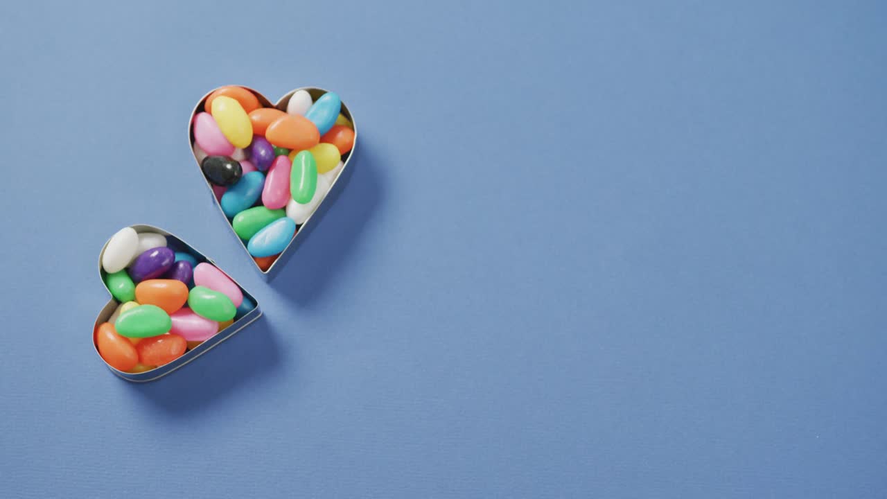 Video of overhead view of multi coloured sweets in two heart shapes with copy space over blue