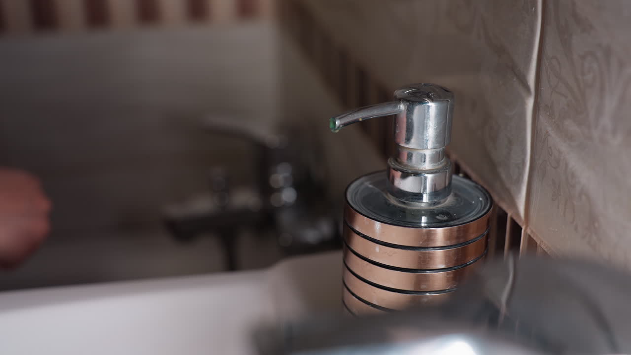Close view fair female hands under soap dispenser tip, one hand ready to receive liquid while other presses pump, chrome container on sink beside tiled wall, hygiene routine, clean bathroom scene