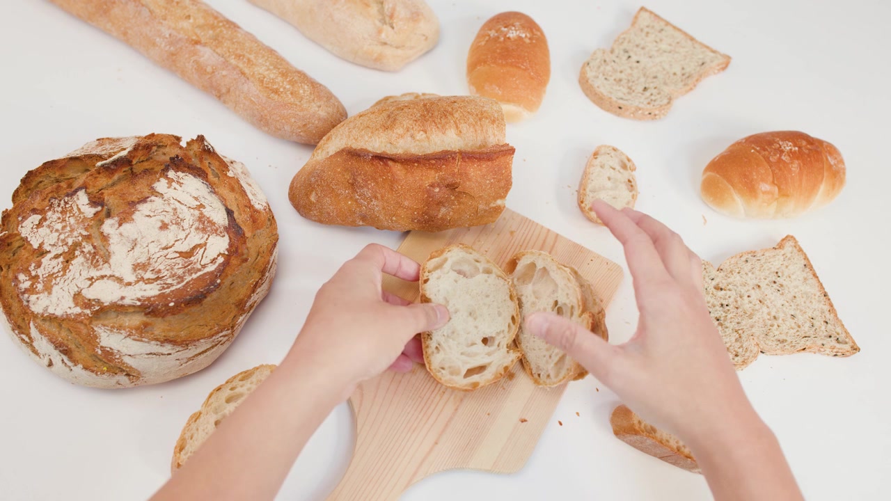 Ripresa dall'alto di mani che sistemano fette di pane a lievitazione naturale fresco su un tagliere, con luce intensa.
