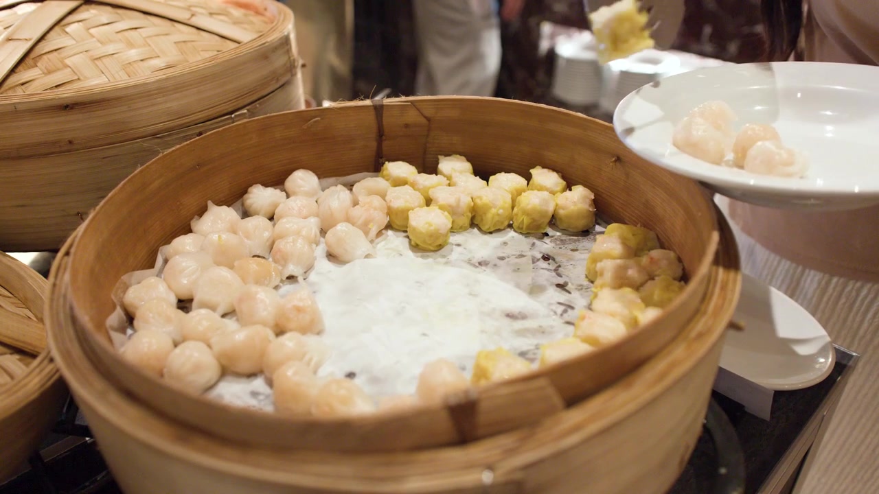 A person uses metal tongs to pick shrimp dumplings from a large bamboo steamer basket
