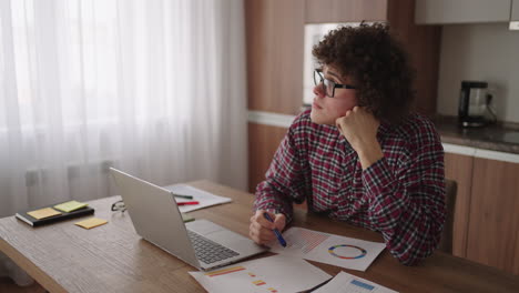 Brooding-serious-curly-freelancer-man-sit-at-table-in-comfortable-home-office-room-work-on-laptop-looks-concentrated-thinking-over-business-issue-solution-makes-telecommute-job