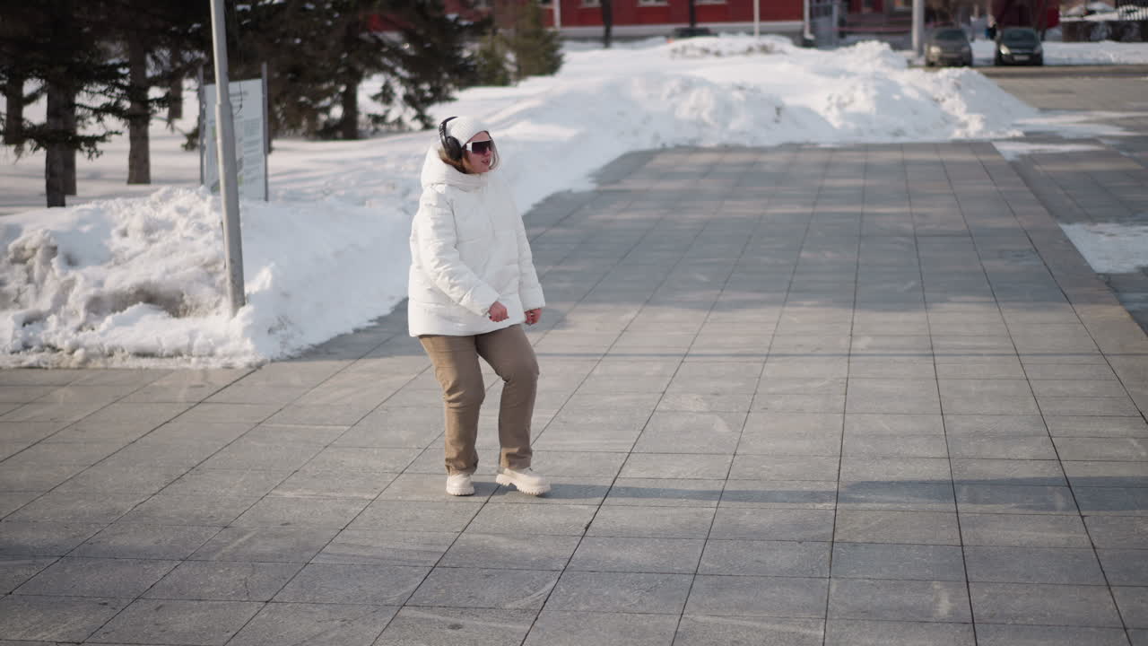 Dance instructor in white winter coat, beanie and headset adjusts dark sunglasses while dancing energetically on paved plaza surrounded by snowbanks, with urban road and trees in background