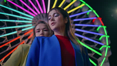 fashion models looking camera at neon illuminated ferris wheel. two girls posing