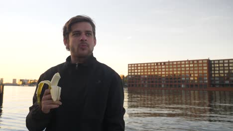 un hombre guapo comiendo un plátano y sonriendo en un muelle durante la puesta de sol.