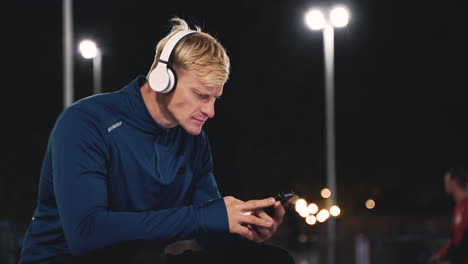 Sportlicher-Blonder-Mann,-Der-Im-Park-Sitzt-Und-Musik-Mit-Bluetooth-kopfhörern-Hört-Und-Ein-Handy-Benutzt,-Während-Er-Nachts-Während-Seiner-Trainingseinheit-Eine-Pause-Macht-1