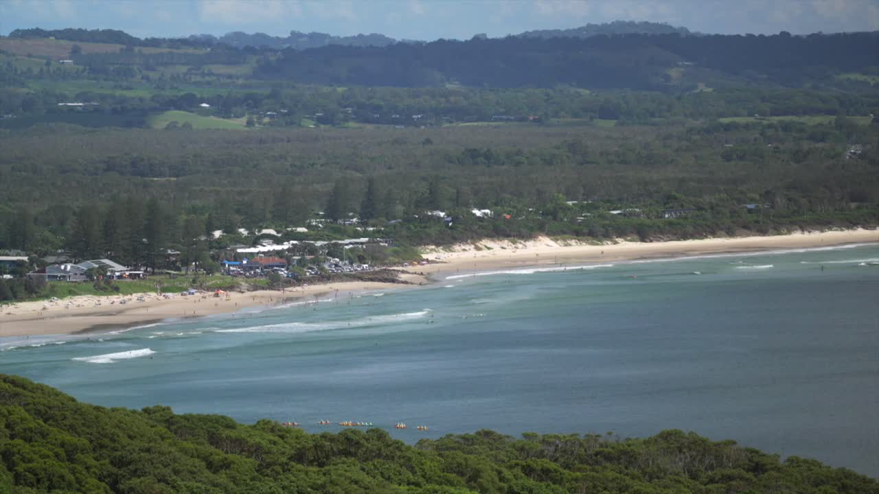 Pemandangan pesisir *long shot* dari pantai berpasir dengan ombak yang pecah di dekat pantai dan hutan pesisir padat yang membentang ke daratan, Byron Bay, Australia