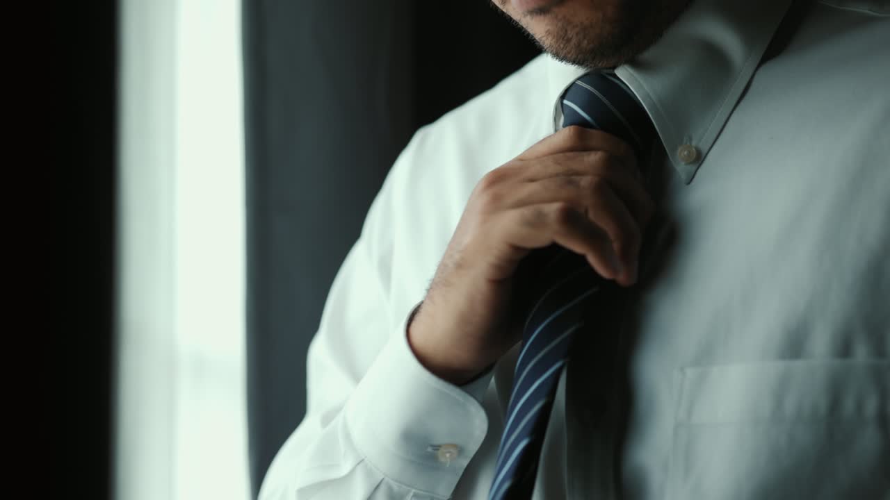 Confident businessman tying or adjust the necktie near window in hotel room in the morning. Handsome man wearing a nice necktie on wedding day.