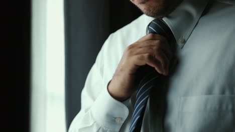 confident businessman tying or adjust the necktie near window in hotel room in the morning. handsome man wearing a nice necktie on wedding day.