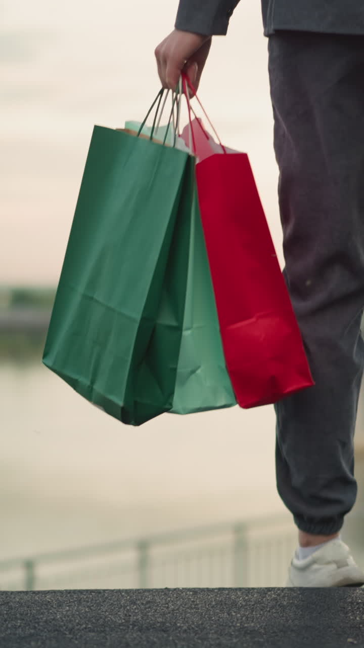 vista trasera de la dama caminando por las escaleras llevando bolsas de compras coloridas, con barandillas de hierro en ambos lados y una alfombra negra bajo los pies, el fondo borroso presenta la orilla del río