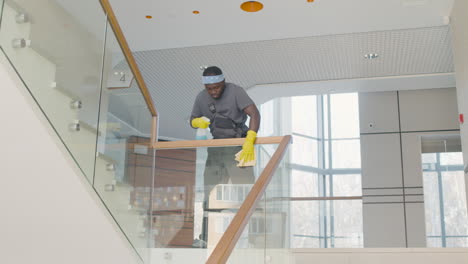 vista inferior del hombre de la limpieza con guantes limpiando la barandilla de la escalera y los cristales dentro de un edificio de oficinas