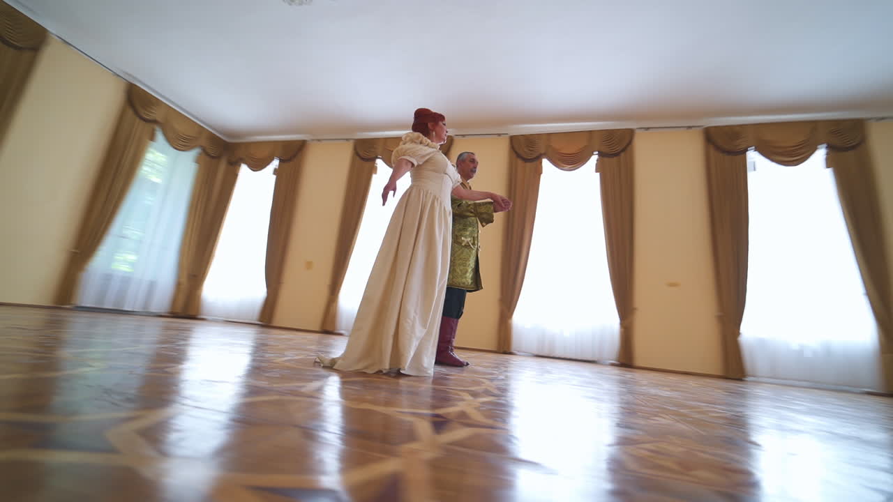 Woman in a long white historic dress dances to a man wearing old golden jacket. Circling footage around the aging couple performing old type dance. Low angle view.