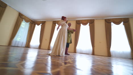 woman in a long white historic dress dances to a man wearing old golden jacket. circling footage around the aging couple performing old type dance. low angle view.