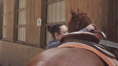 close up over the back of a cowgirl putting a saddle on a brown red horse western style in a dusty warm arena 4k in south eastern michigan