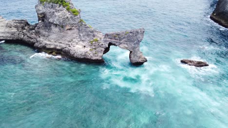 Waves-breaking-into-white-foam-on-Natural-arch-Bridge-against-Atuh-Beach,-Nusa-Penida-Island