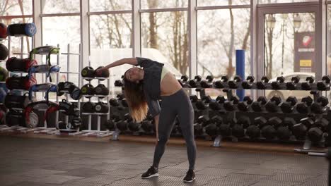 Full-length-shot-of-determined-fitness-woman-warming-up-in-modern-gym.-Muscular-sportswoman-warming-up-before-a-intense-workout-standing-in-front-the-wall-of-dumbbells-and-bright-window