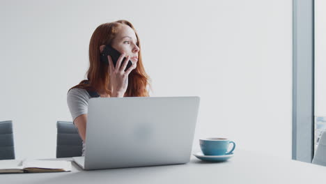 casually dressed young businesswoman working on laptop in modern office taking call on mobile phone