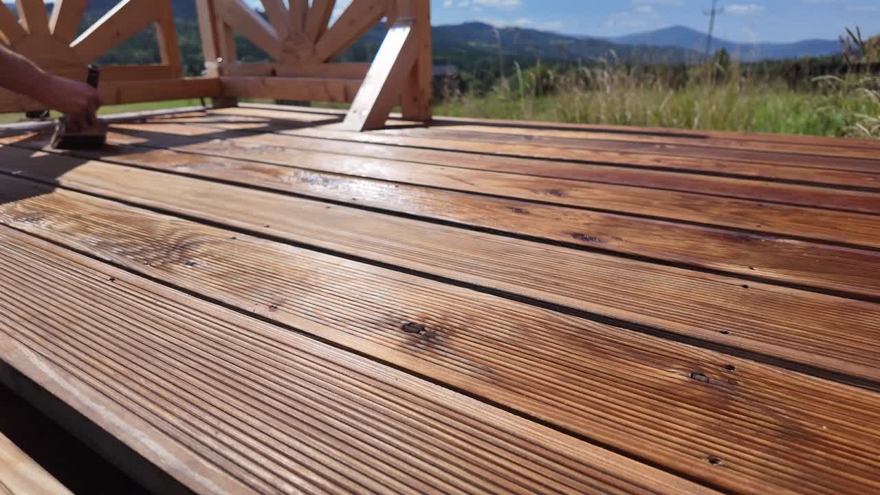 Working man oils varnishing impregnating wooden terrace boards. Decorative railing, serene sky, clouds, and mountain horizon evoke home improvement and outdoor maintenance themes - DIY work
