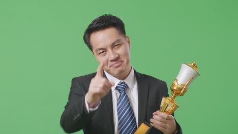 close up of asian business man in a suit and tie with a gold medal and trophy disapproving with no index finger sign and smiling to camera on green screen background in the studio