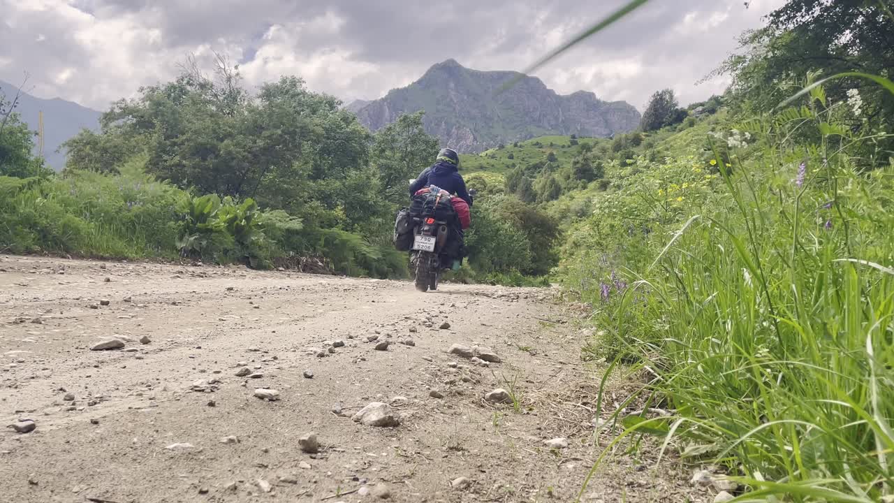 Vista posteriore di una moto da avventura a pieno carico che percorre una strada di montagna sterrata in Kirghizistan, mostrando viaggi fuoristrada, bagagli pesanti e un lussureggiante paesaggio di valle alpina verde