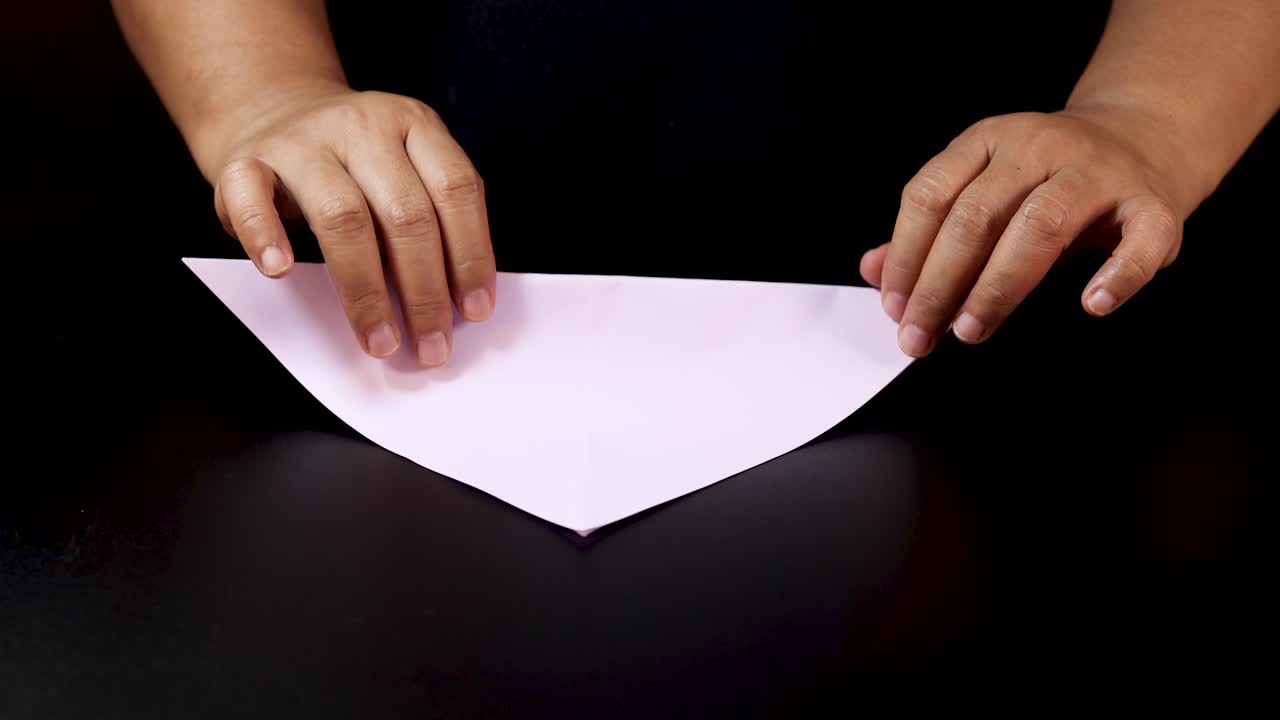 Adult hands carefully fold purple paper on black surface under soft, even lighting, overhead view