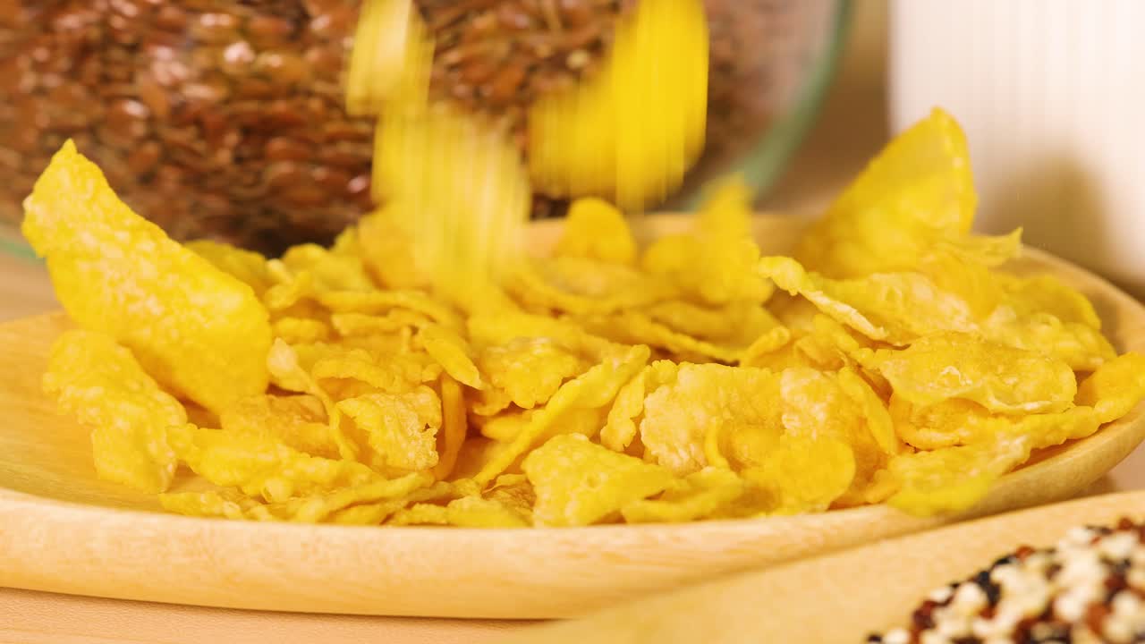 Cornflakes are poured into a bowl, surrounded by grains. Bright lighting enhances the golden color and texture