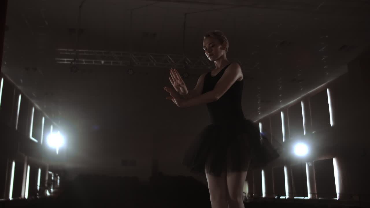 Graceful woman ballerina in a dark dress on a dark stage of the theater in the smoke performs dance moves in slow motion.