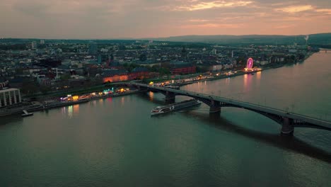 Toma-Aérea-De-Dron-De-Mainz-Con-Un-Barco-De-Cruce-Y-El-Puente-Principal-Del-Río-Rin-Bajo-Una-Puesta-De-Sol-De-Verano-Naranja-Con-Muchos-Reflejos-En-El-Agua-Y-Un-Festival-De-Vino-En-La-Parte-De-Atrás