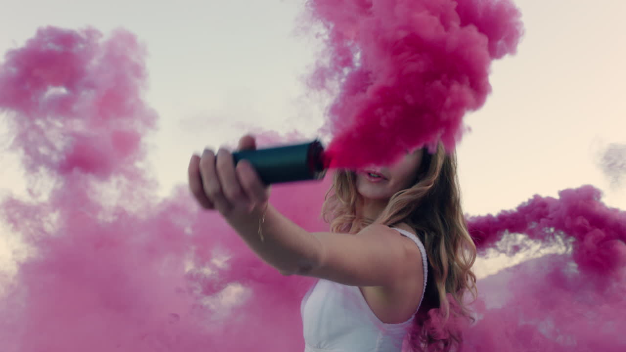 bellissima donna che agita una bomba di fumo rosa che balla sulla spiaggia all'alba celebrando la libertà creativa