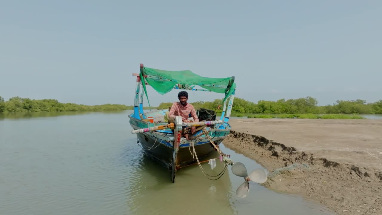 A solitary fisherman sits on the bow of a traditional boat, navigating the calm waters of the Indus River in Karachi, surrounded by lush greenery and a serene atmosphere.