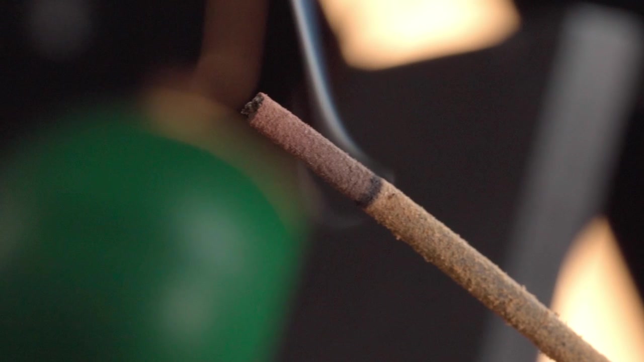 Scatto macro ravvicinato di un bastoncino d'incenso che brucia e rilascia fumo su uno sfondo scuro sfocato, evidenziando il legno texturizzato, la punta incandescente e i dettagli atmosferici