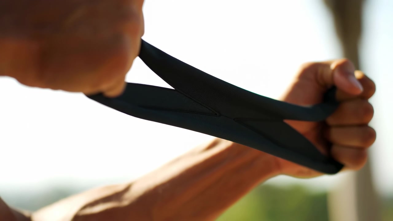 Two hands stretch black elastic strap with visible tension, slight deformation, strong grip, forearm strain, bright backlight, warm sunlight, soft blur, and outdoor beach atmosphere