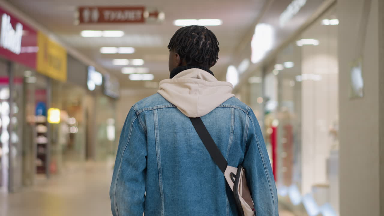 young man walking through modern shopping mall corridor wearing denim jacket hoodie and crossbody bag capturing urban lifestyle exploration with blurred storefronts and bright ambient lighting