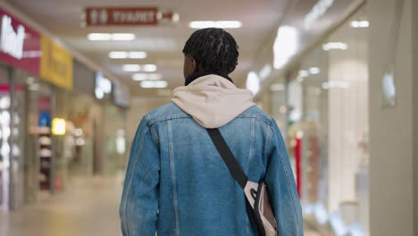 young man walking through modern shopping mall corridor wearing denim jacket hoodie and crossbody bag capturing urban lifestyle exploration with blurred storefronts and bright ambient lighting