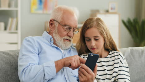 Porträt-Des-Süßen-Blonden-Teenager-Mädchens-Und-Seines-Grauhaarigen-Opas,-Der-Ihr-Etwas-Auf-Dem-Smartphone-Bildschirm-Zeigt,-Der-Zu-Hause-Auf-Der-Couch-Sitzt