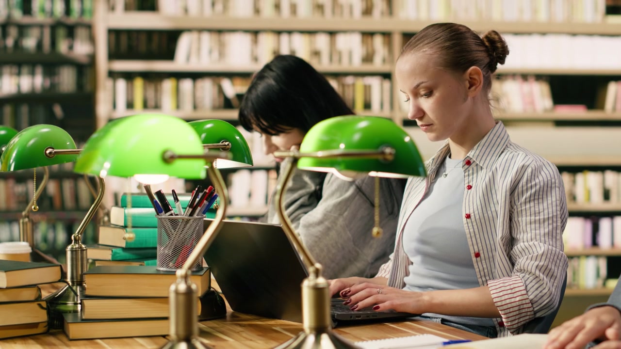 Studente che digita sul laptop e prende appunti da un libro aperto alla scrivania della biblioteca.