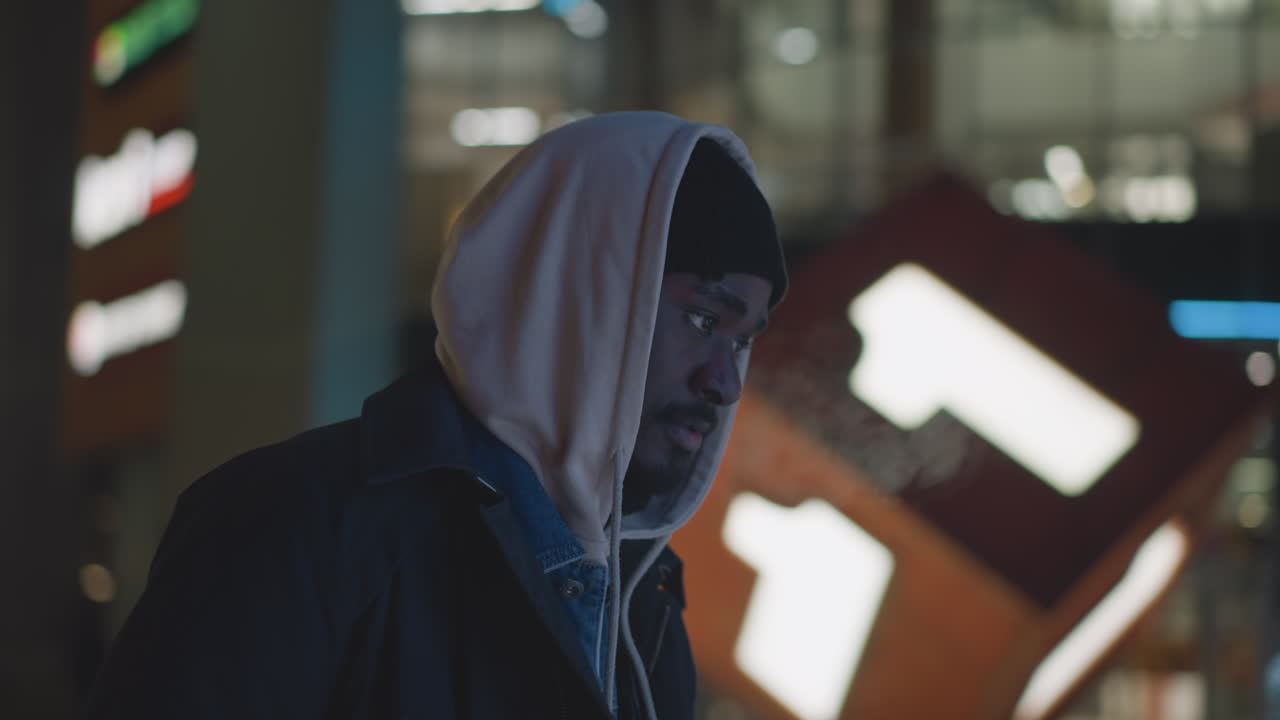 Man standing outside at night, looking down with glowing number-shaped decoration in background. Urban setting with street lights and illuminated signs, creating a festive, holiday atmosphere